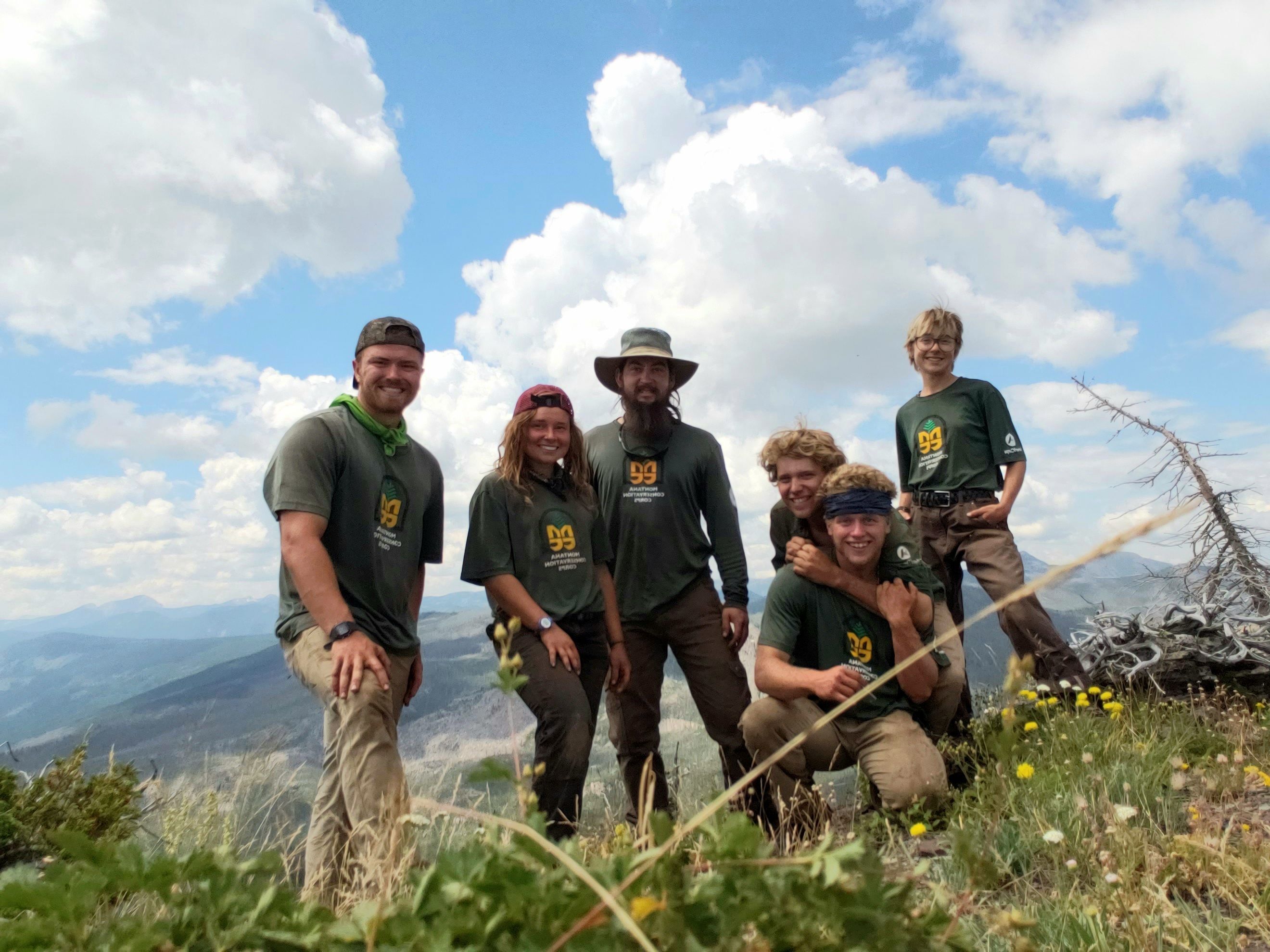 A happy crew stands on a mountain summit. There are wildflowers and grass in the foreground and a blue sky in the background.