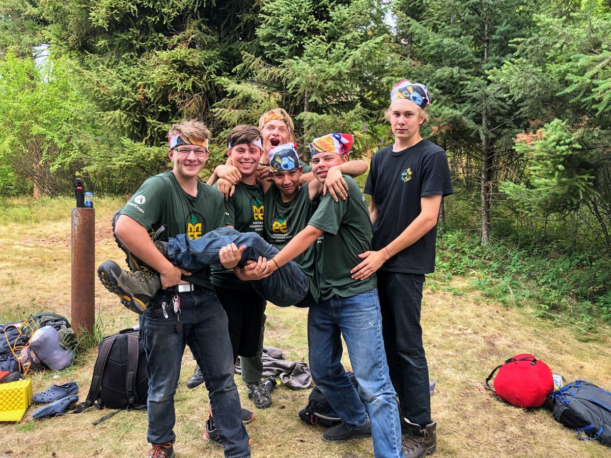 [Image Description: Six MCC youth members are standing in their campsite, joyously hoisting another member up into the air, all smiling and laughing.] 