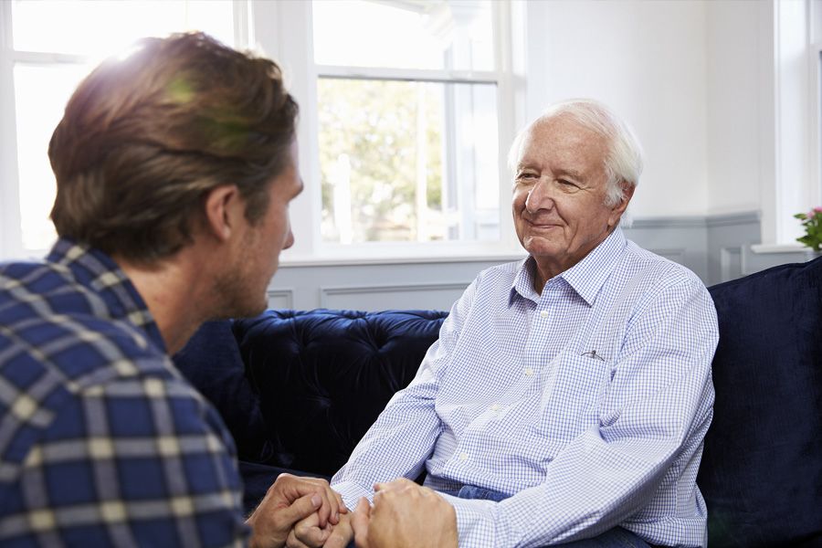 Older man smiling gently as he sits on his couch. A young man sits across from him and they're clasping hands.