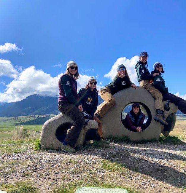 The women's fuels and fire crew sits on cement sculptures. with a bright blue sky and rolling hills in the background.