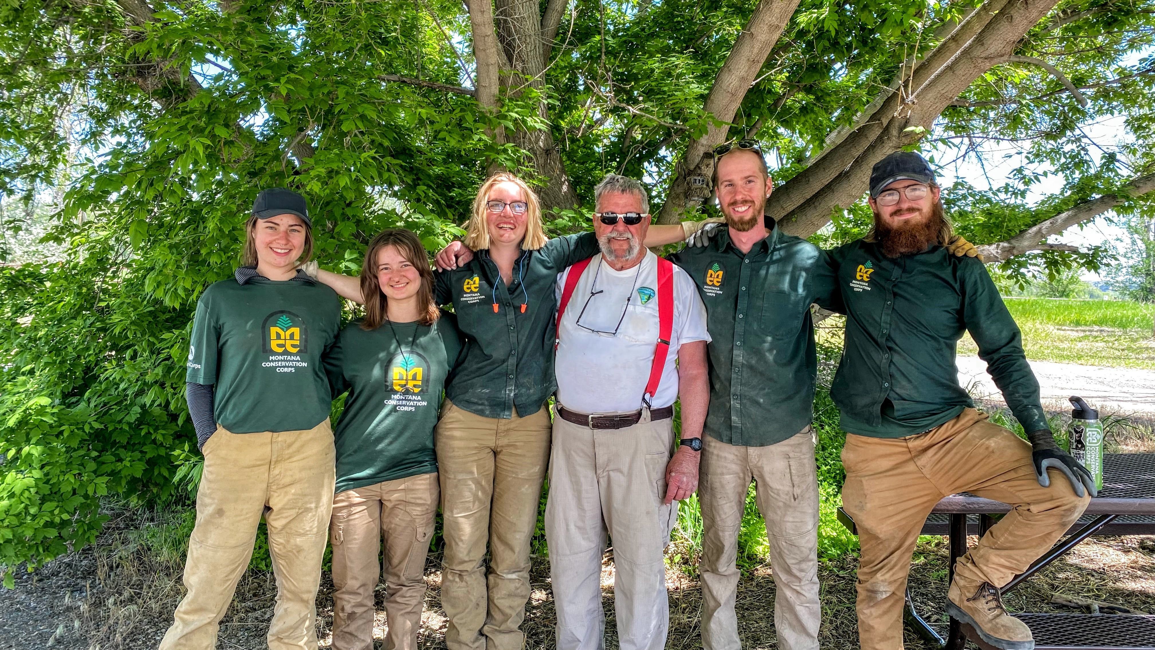 A group stands in front of a tree. In the center is an older man wearing a white shirt and red suspenders, this is Don the BLM Billings project partner.
