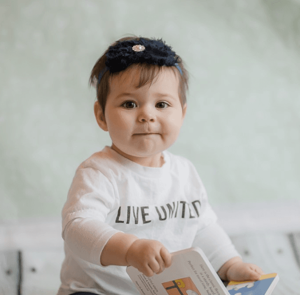 a baby with dark hair and a book wearing a Live United shirt