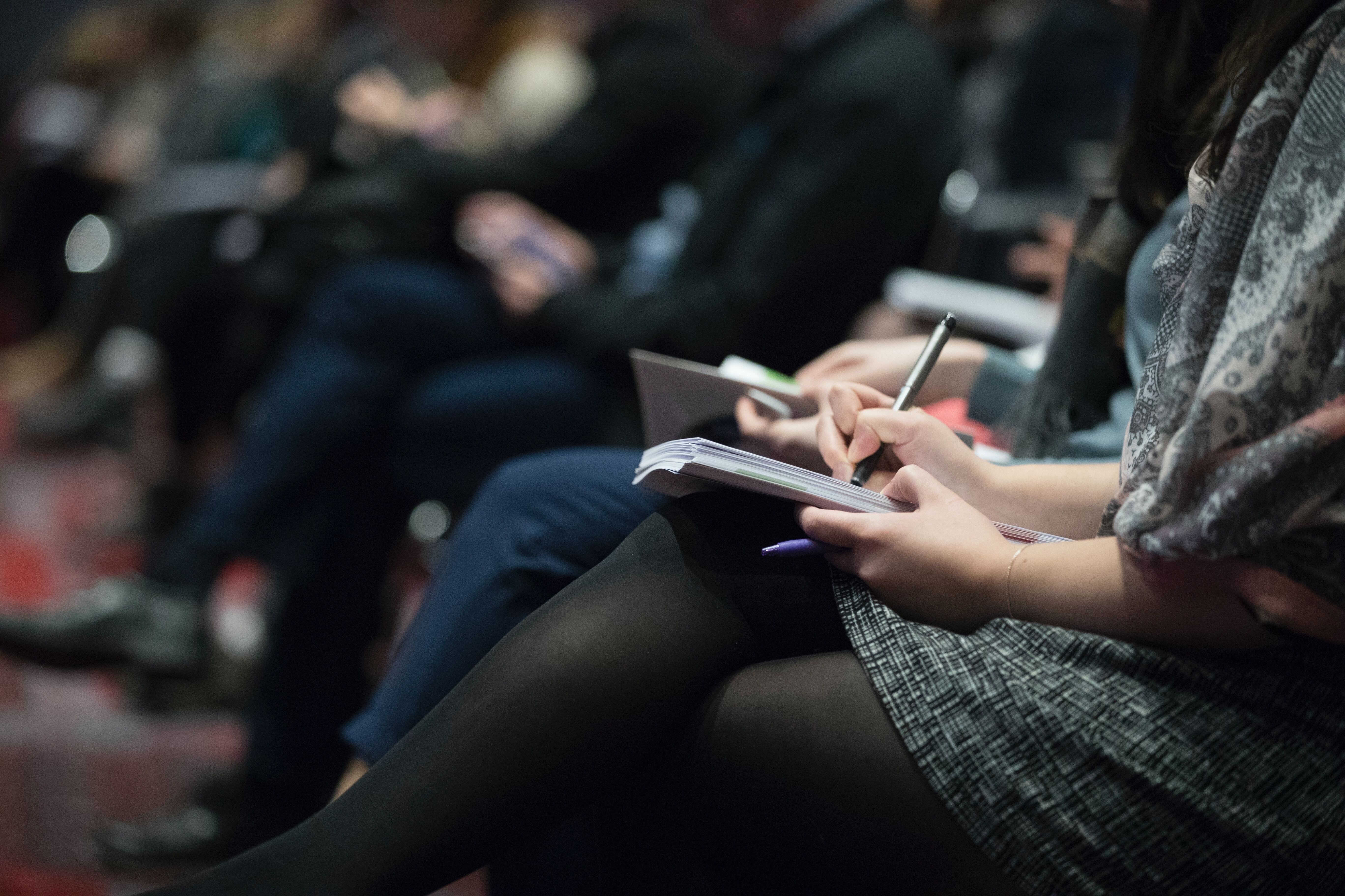 Picture of a row of people sat at a conference, only their laps are visible and one person is writing in a notebook