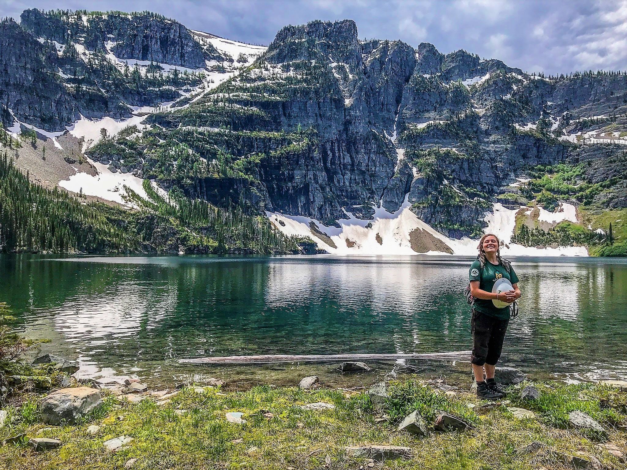 [Image Description: One MCC member stands in front of a turquoise lake, with a peak in the background, covered in a dusting of snow. The member is standing on the edge of the lake in their uniform, holding their hard hat.] 