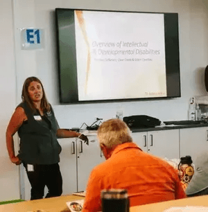 A woman is presenting in a classroom setting next to a screen displaying a slide titled Overview of Intellectual and Developmental Disabilities. Attendees are seated at tables with documents and water bottles. The environment appears educational and profe