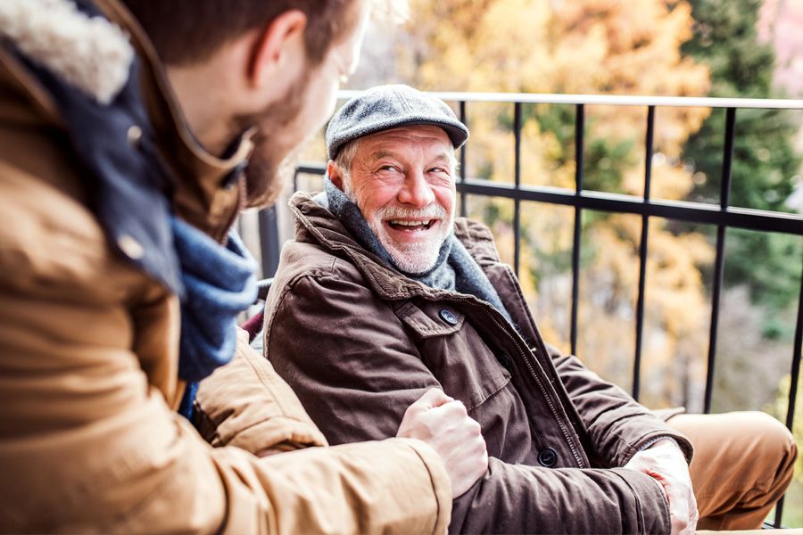 Older man sitting in wheelchair on a balcony, smiling at his son who is out of focus