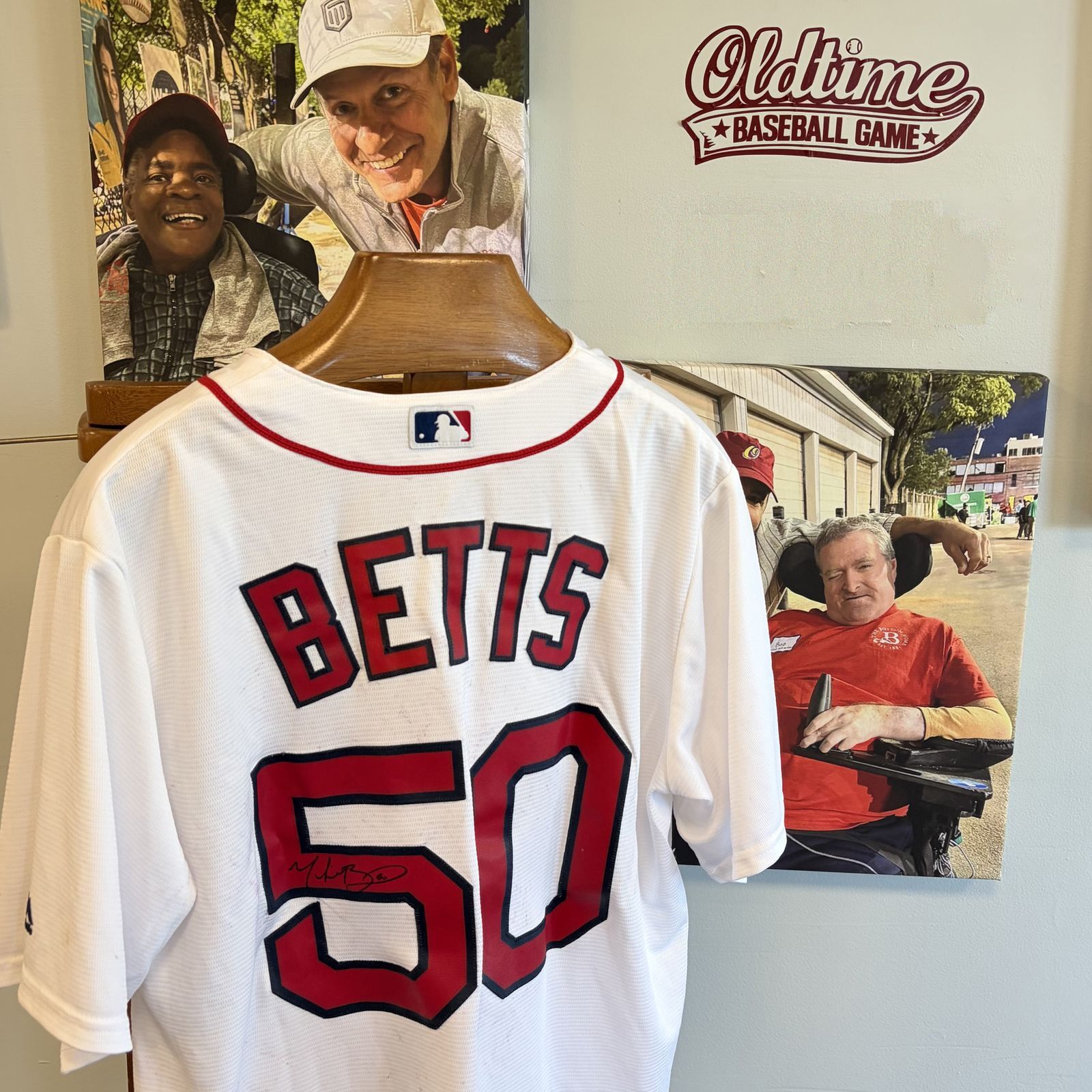 A white Boston Red Sox jersey signed by Mookie Betts hangs on a wooden hanger in front of a display celebrating an Oldtime Baseball Game. The back of the jersey reads "BETTS" above the number "50" in red with navy trim. Behind the jersey are two large pho