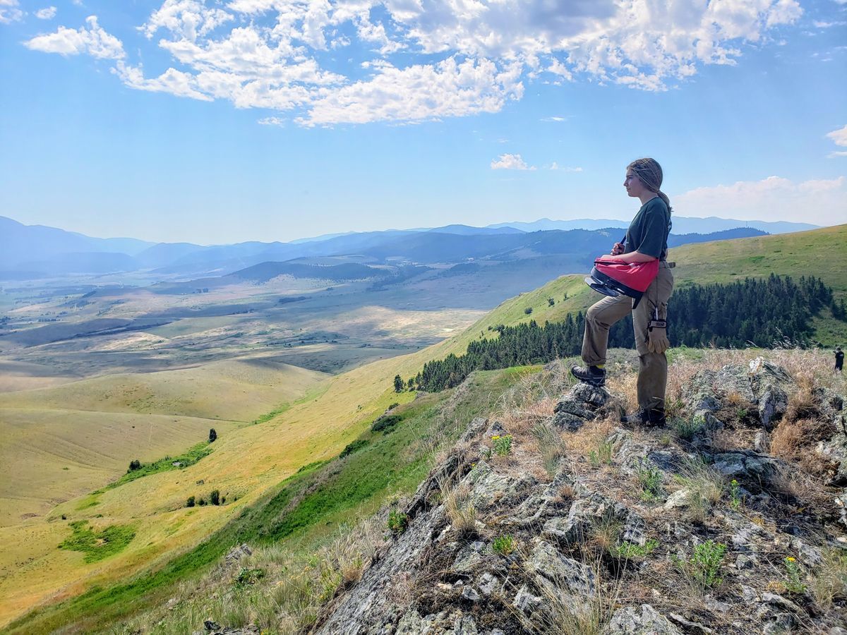 [Image Description: An MCC youth member standing on the edge of a rocky mountain, staring into the golden valley far below them. Blue mountains paint the edge of the the image, and white clouds pepper the sky above them.]