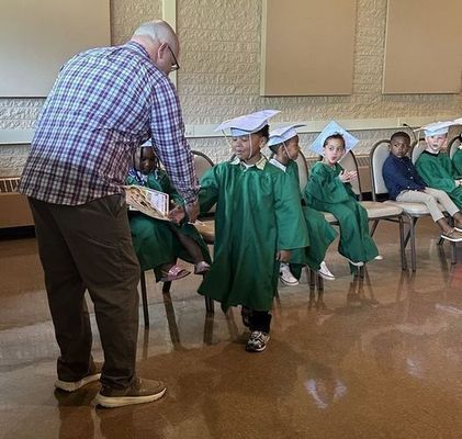 A preschooler, wearing a green cap and gown, shakes hands with a man while recieving a piece of paper. Other preschoolers watch, dressed the same.