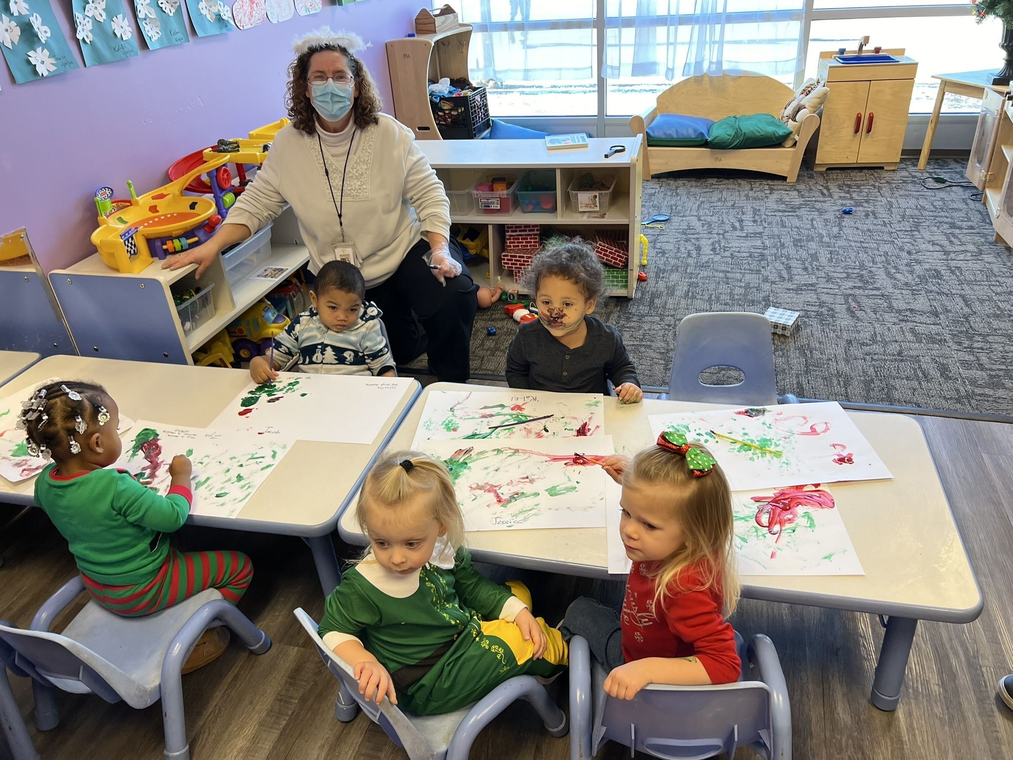 A teacher wearing a mask sits at a dask in a classroom with a few other students. The students all have paintings in front of them they are working on.