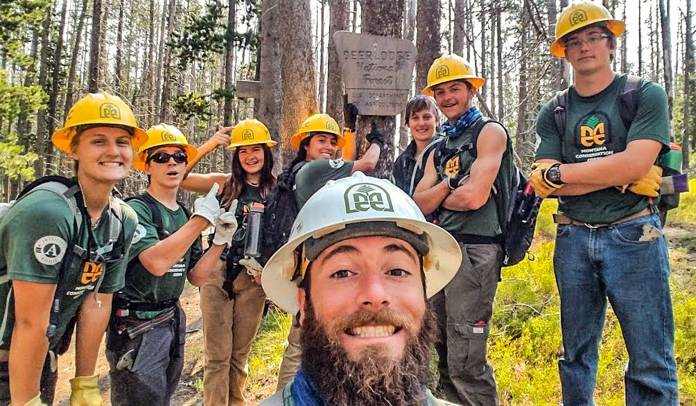 [Image Description: Eight MCC Crew members and their leader standing in front of a USFS border sign that states 'Deerlodge National Forest'. ]