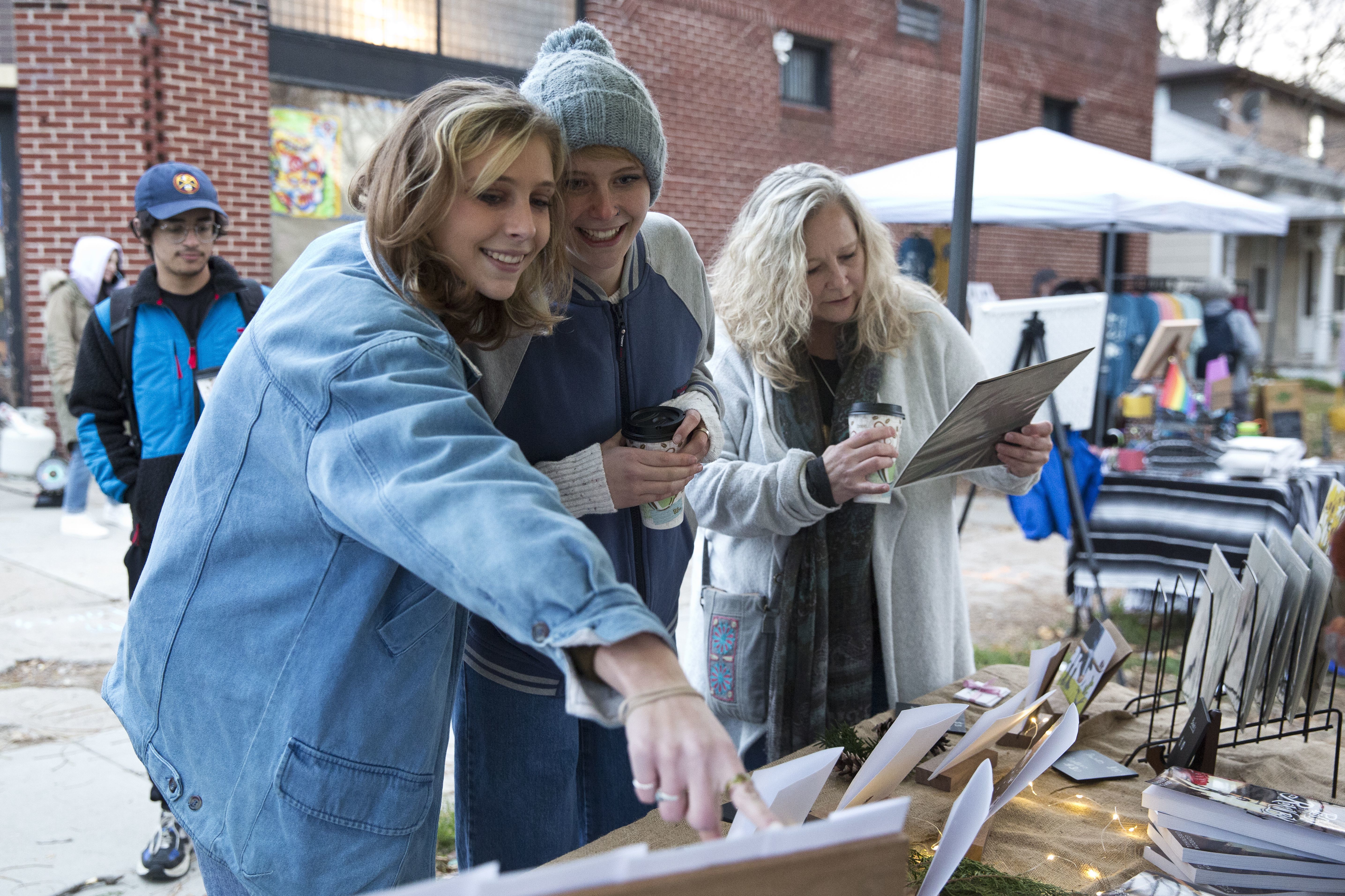 Three smiling neighbors pointing to products on a table at the Holiday Makers Market. 