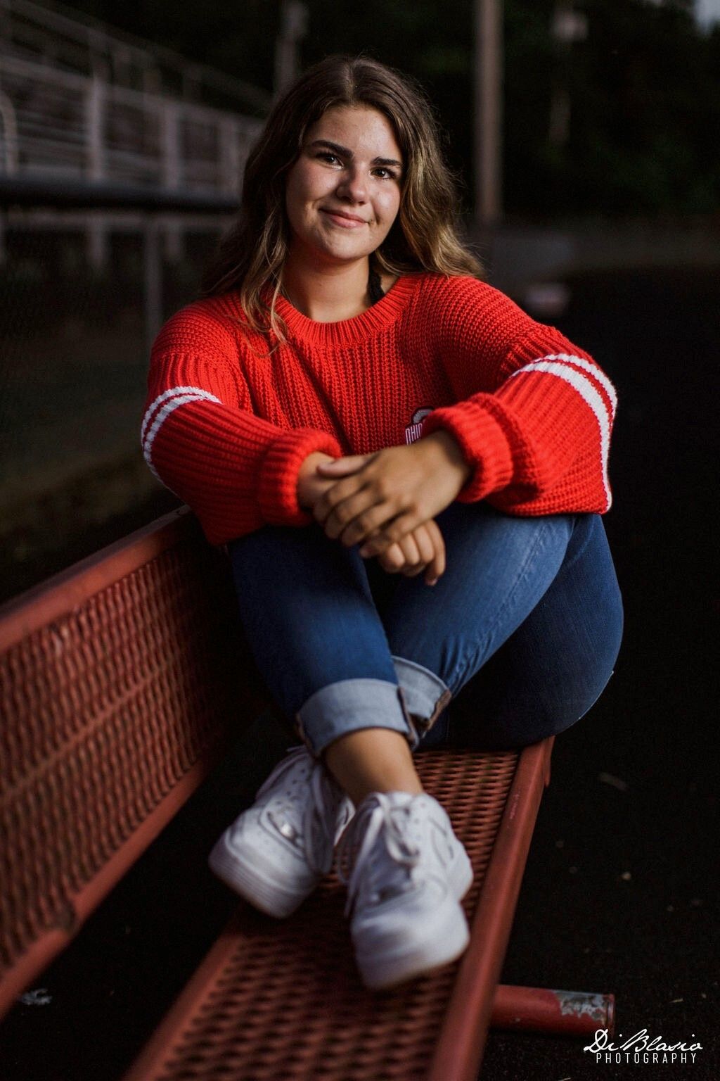 Mya Lepley happily sits on a red bench with her arms wrapped around her crossed legs.