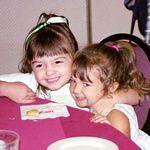 Two young girls with their arms around each other posing for a picture at a table covered with red table cloth. 