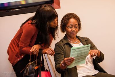 A woman sitting and pointing to something on a green piece of paper. Behind her is another woman leaning over the chair looking at the paper.