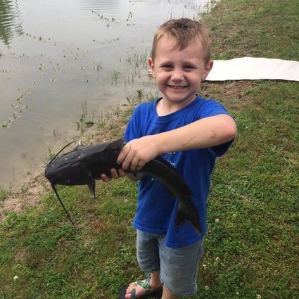 Smiling young boy in a blue shirt and shorts holding a freshly caught catfish near the edge of a pond on a grassy bank