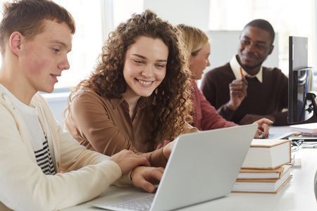 A photo of 4 college students who sit at a table. They are paired off in groups of 2, each group looking at a separate computer.
