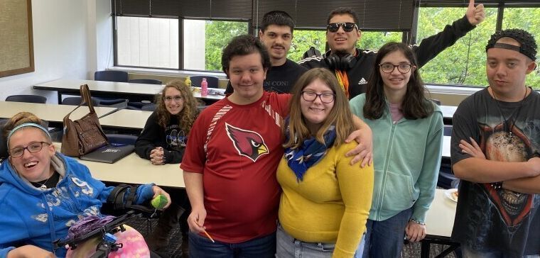 8 students with ID pose for a photo in a classroom. One is in a wheelchair, one sits at a desk, and the rest stand together. They all smile for the camera.