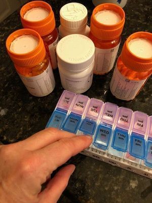 A woman's hand can be seen pulling pills out of a colorful pill box with days of the week written it. Additional pill bottles can be seen behind.