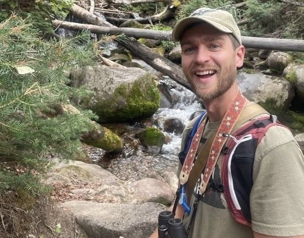 Tobin Brown smiles near a stream in the woods. He is wearing binoculars, a baseball cap, and a backpack.