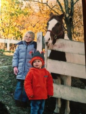 Photo of Bella in a big blue coat, Jack in a bright red coat, with a brown and white horse on the other side of the fence behind them.