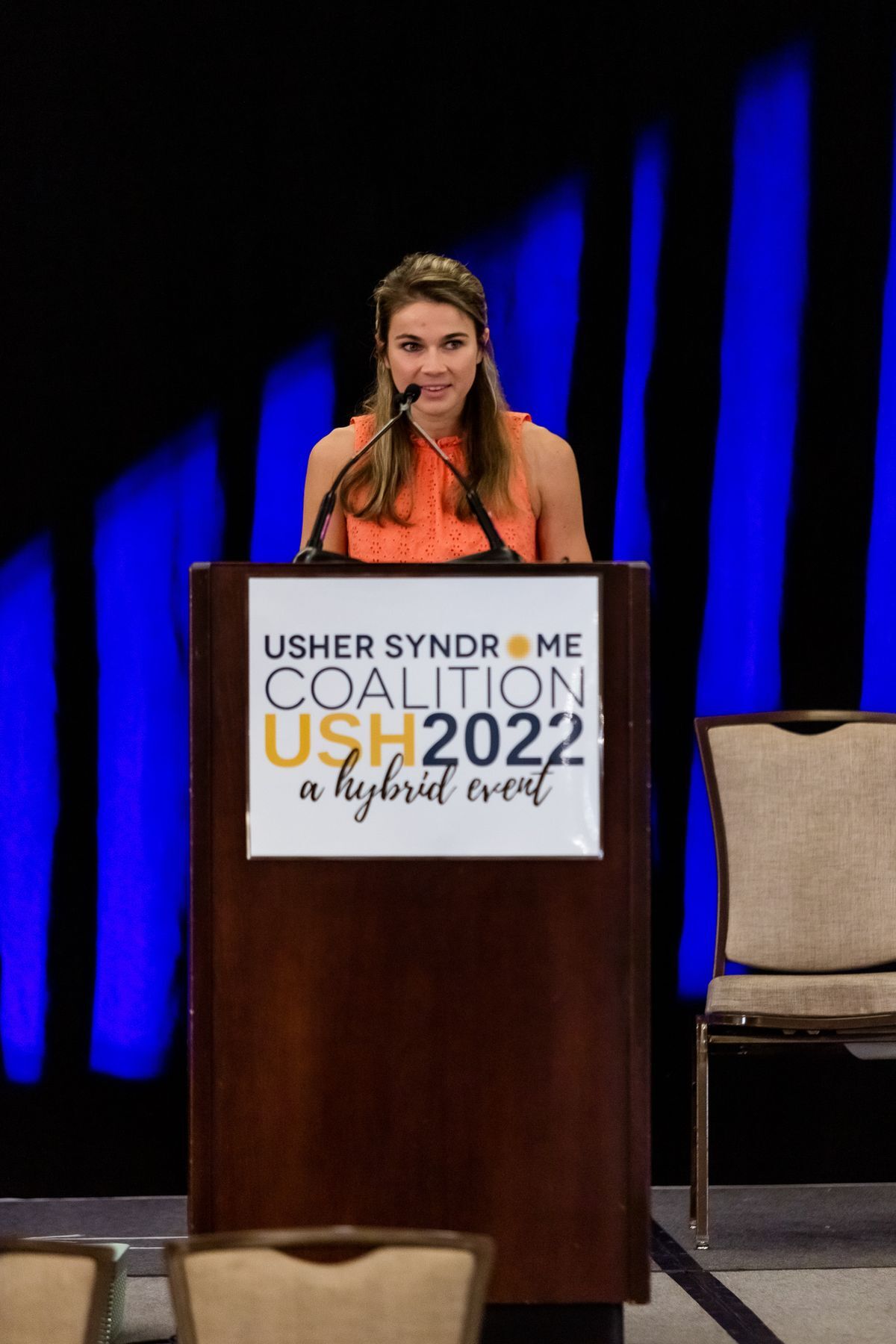 Becca Meyers standing at the podium wearing an orange shirt, talking.