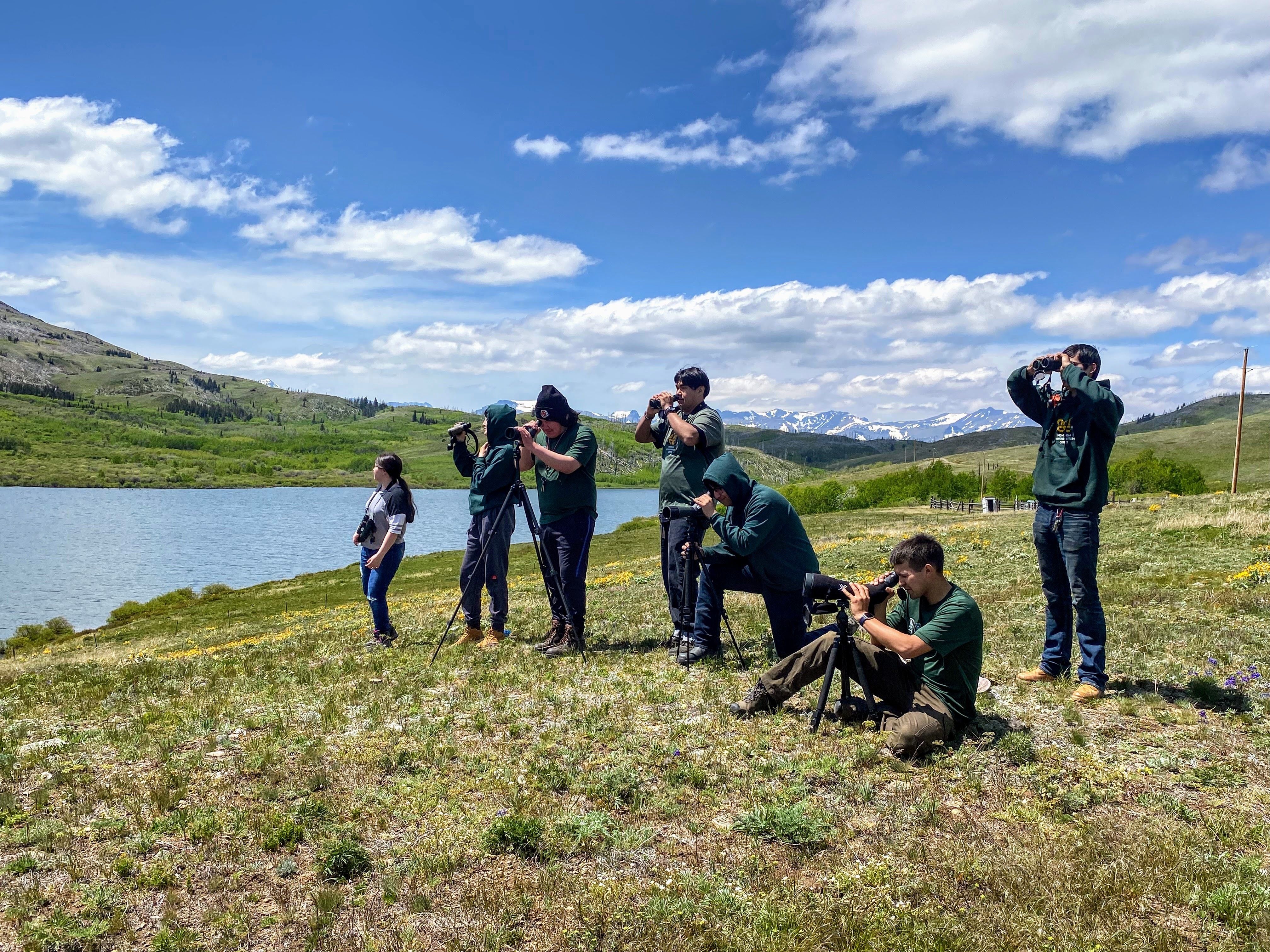 [Image Description: Seven members of the Piikuni Lands Crew stand in a field next to the river, looking through binoculars.]