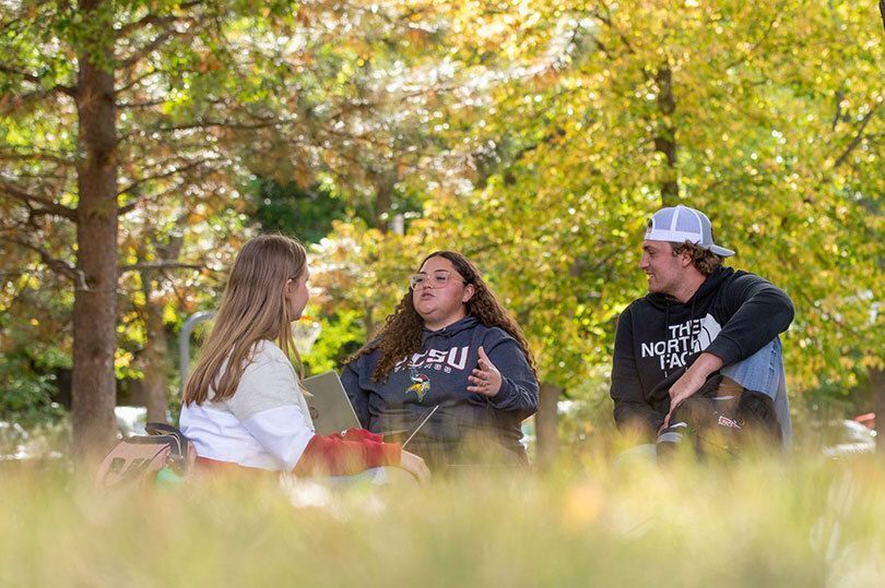 Three students sitting on the VCSU front lawn, engaging in conversation, surrounded by fall-colored trees
