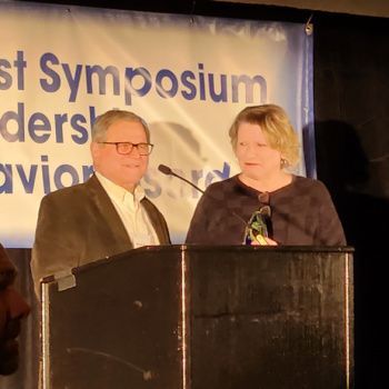 At the Midwest Symposium for Leadership in Behavior Disorders, a bespectacled man in a suit and a short-haired blonde woman in a dark top stand behind a black podium. The woman holds a translucent award trophy as they address the audience.