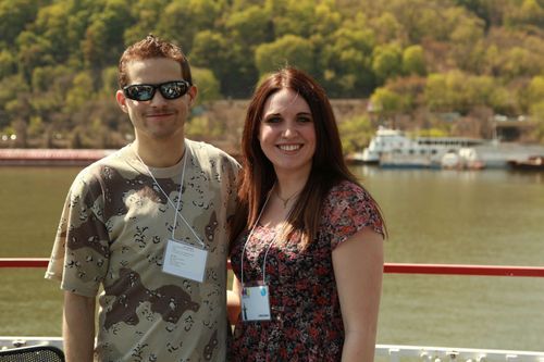 A man and woman, both smiling, stand next to one another. They appear to be on a boat. Water is behind them, and there is a white boat far in the background.