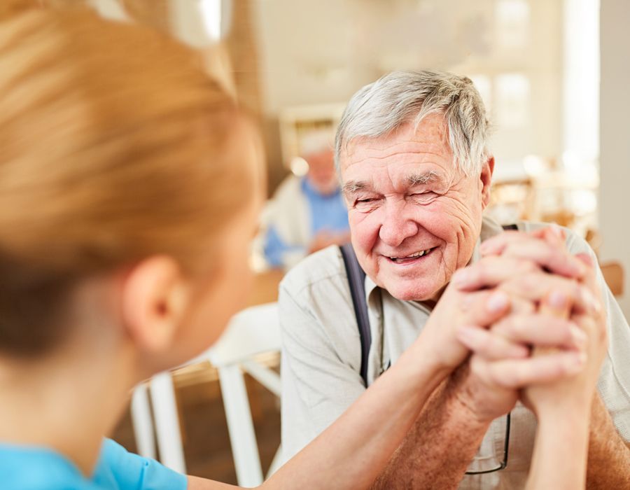 Smiling older man sitting with his caregiver in an assisted living home. They're clasping hands.