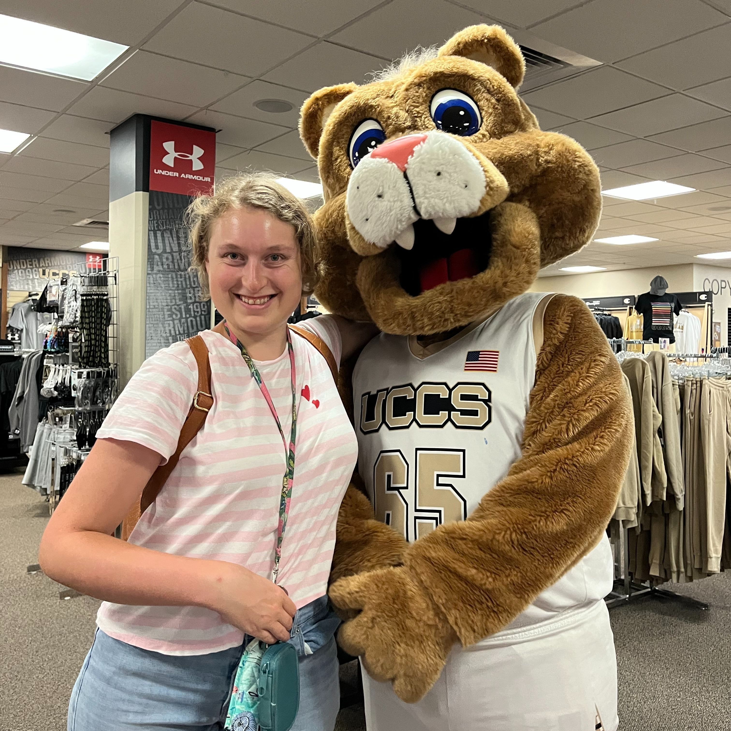 Makena wears a white and pink striped shirt. She stands with the UCCS mountain lion mascot and smiles.