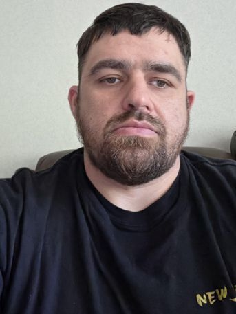 White man with dark brown hair and beard and dark shirt looks at the camera