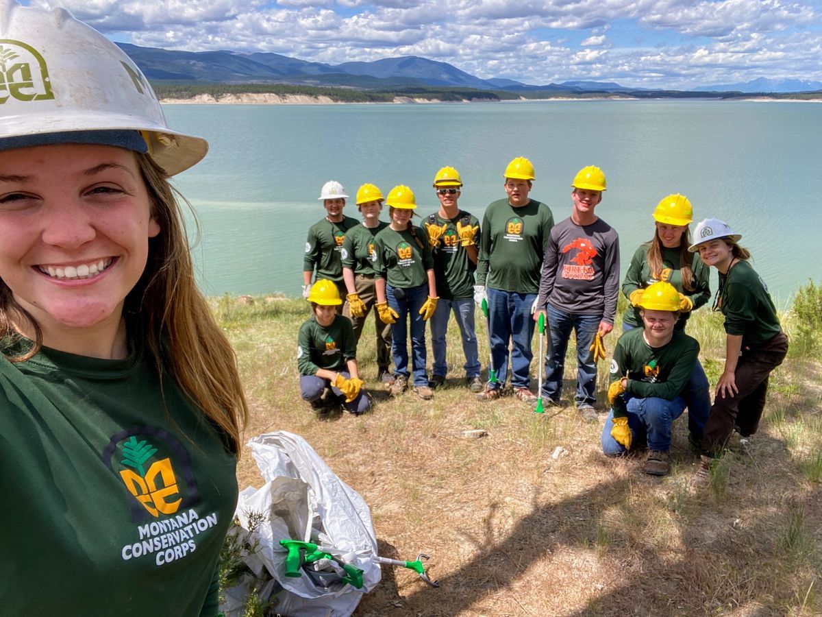 [Image Description: A group of MCC youth members, all grouped together for a selfie on the shore of a large blue lake, sporting hard hats and gloves.] 