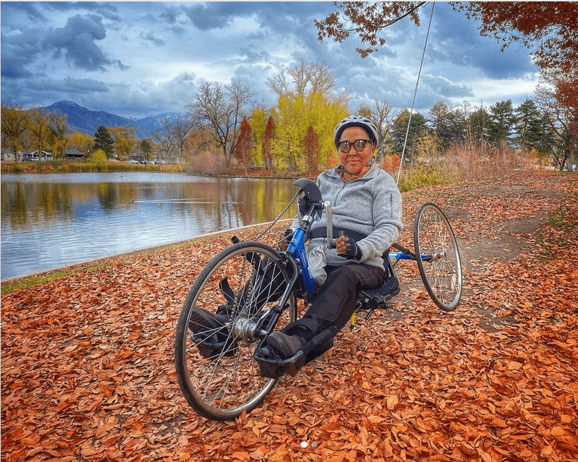 Person sitting on a hand-cycle in leaves next to a lake with mountains in the background