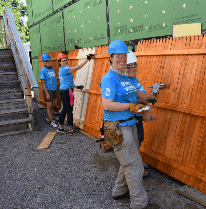 Volunteer smiles while wearing a blue Habitat for Humanity hard hat. She also has a pencil in her ear. 