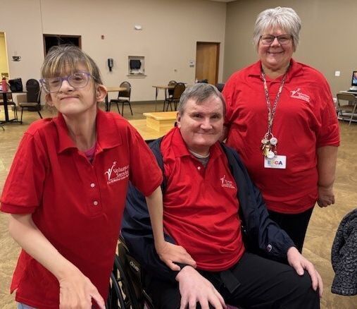Three people face the camera, all wearing red polo shirts with the VA logo on them: a young woman, a man in a wheelchair, and an older woman.