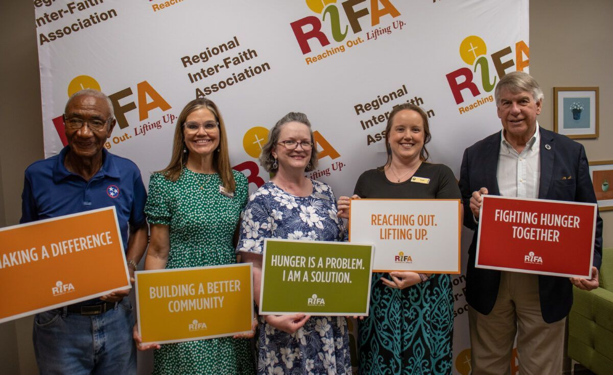 Five people holding signs with messages about community and hunger relief in front of a Regional Inter-Faith Association backdrop.