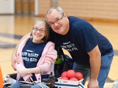 photo of a young woman in a power chair with her dad in a blue t-shirt with his arm around her.