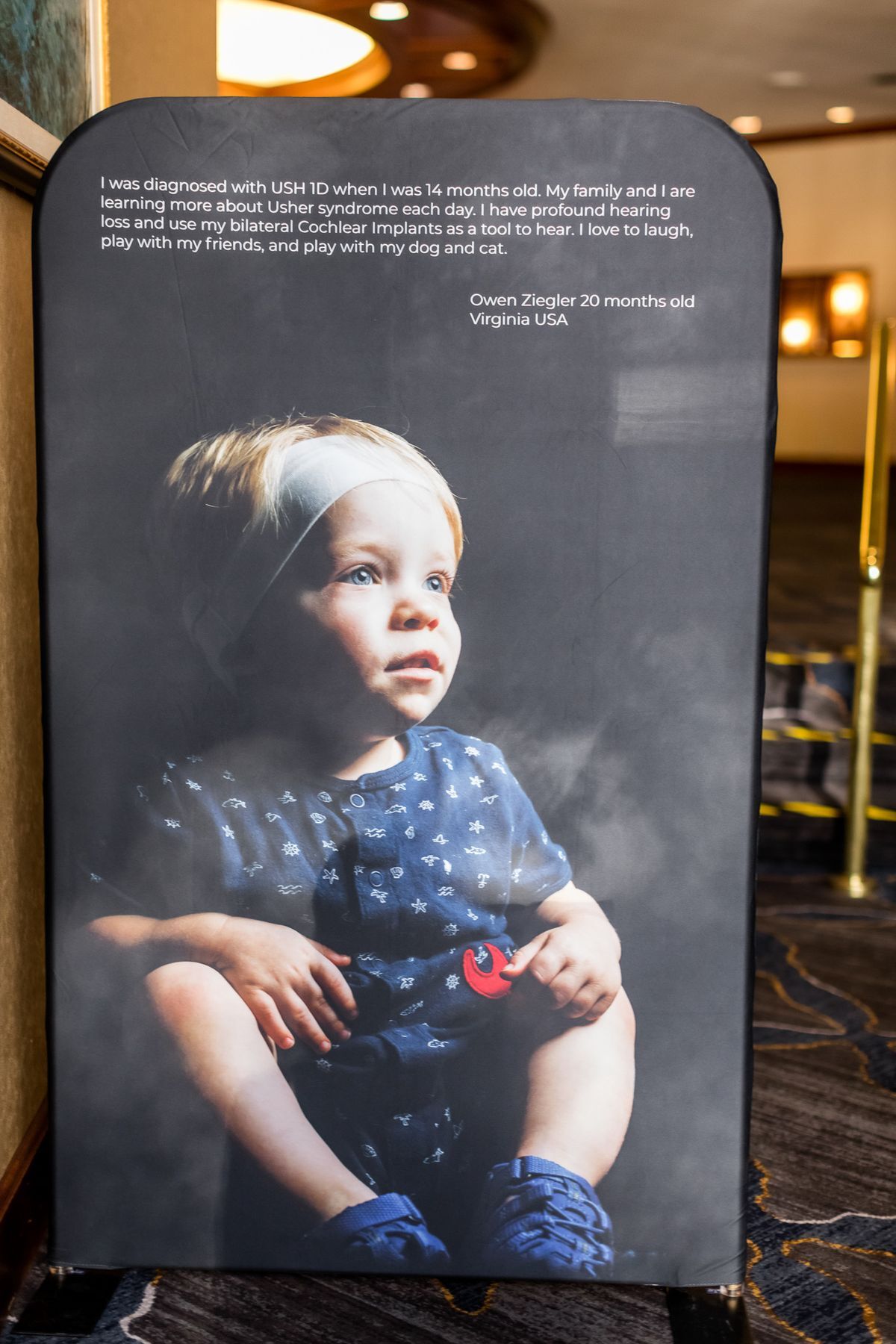 One of the portraits provided by USH Society displayed in the hallway. This one is of an small child in a blue polka dot outfit. She's looking up, away from the camera. In very small print is her story above her photo.