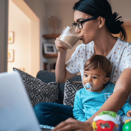 Woman sipping coffee in front of a laptop with a baby on her lap