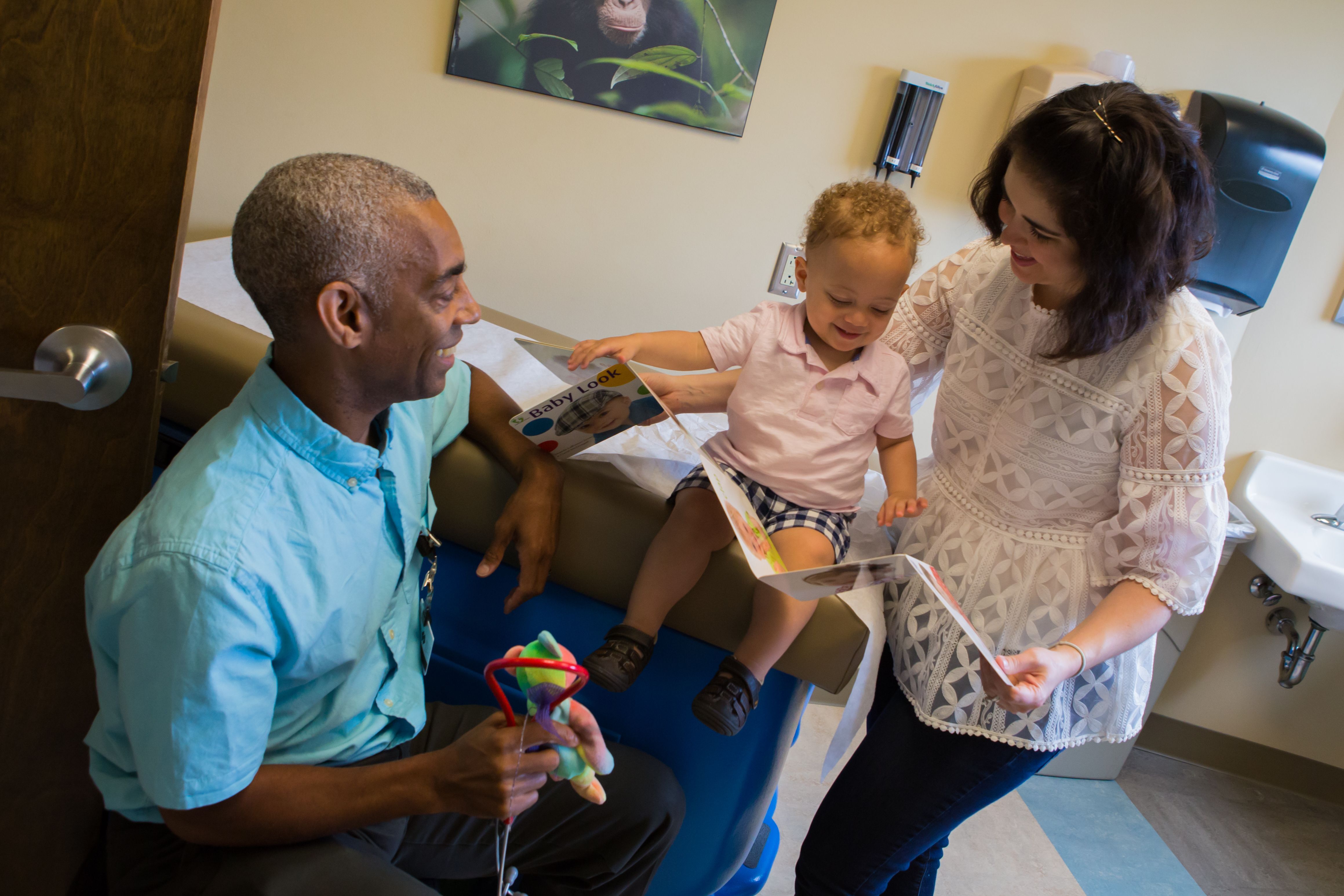 A doctor holds a toy and smiles at a baby looking at a book with his mother.