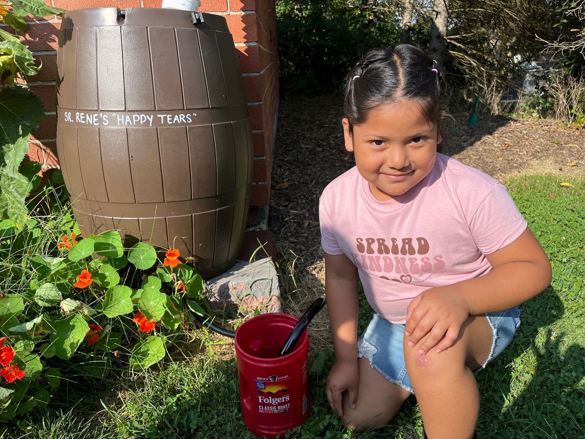 A young girl kneels next to a rain barrel with the words "Sr. Rene's Happy Tears" painted on it. 