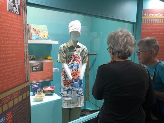 Two women standing in front of an exhibit case at the Smoky Hill Museum showing a mannequin dressed in 1950s male clothing and grilling cooking apron.