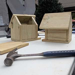 Two finished wooden birdhouses on a table next to a hammer