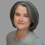 Portrait of a woman with short, gray hair, wearing a light gray shirt. She is smiling slightly and standing against a plain gray background.