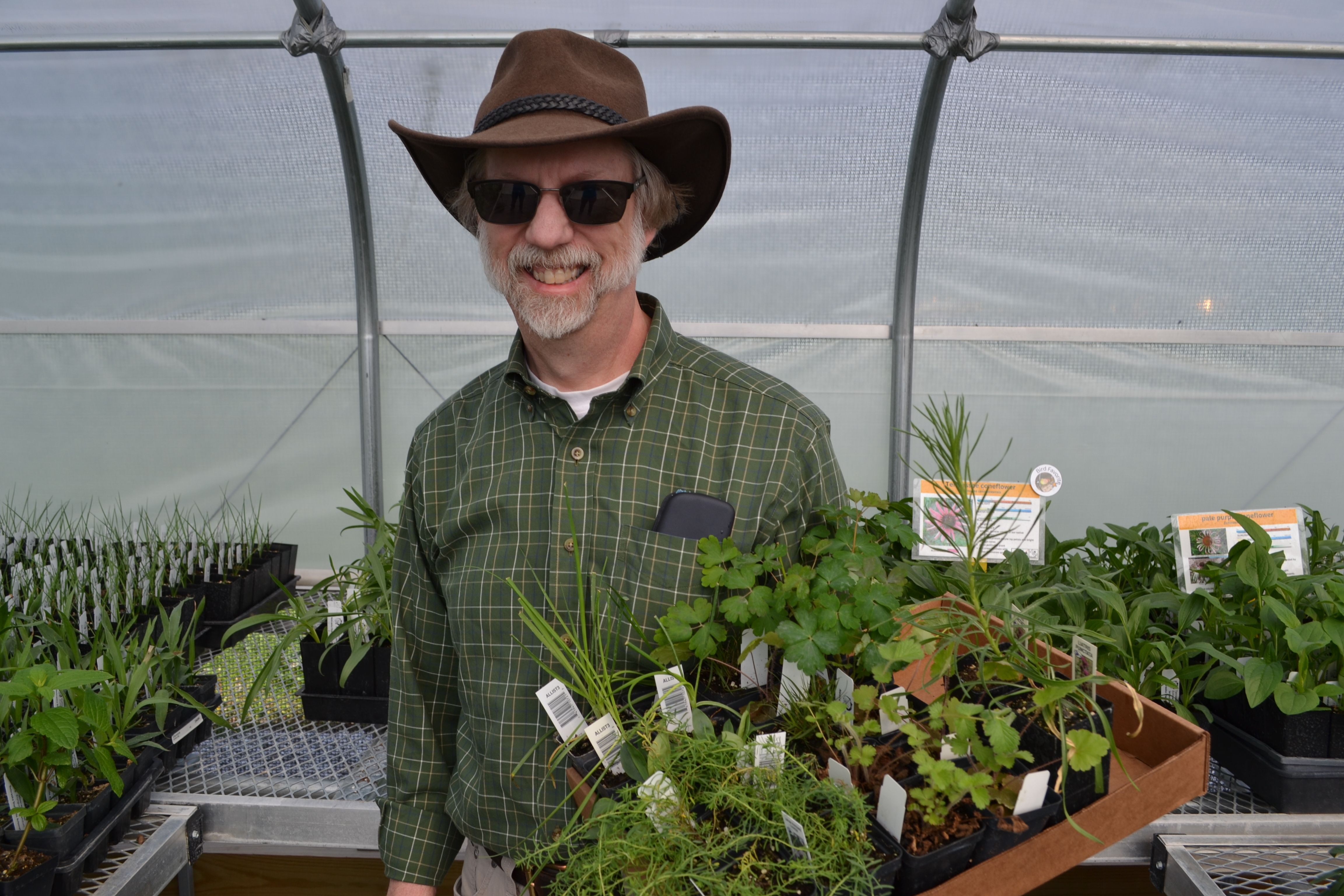 Man wearing a brown cowboy hat and sunglasses holds a box of plants in a greenhouse. 