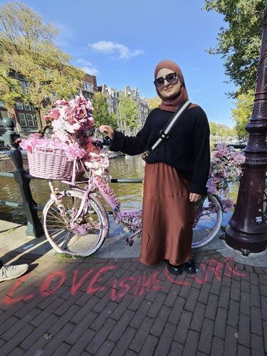 Young woman stands on a street  in front of a river. Her hand touches a basket  full of pink flowers on a pink bicycle. She is wearing a brown hijab, black sweater, and long brown dress.