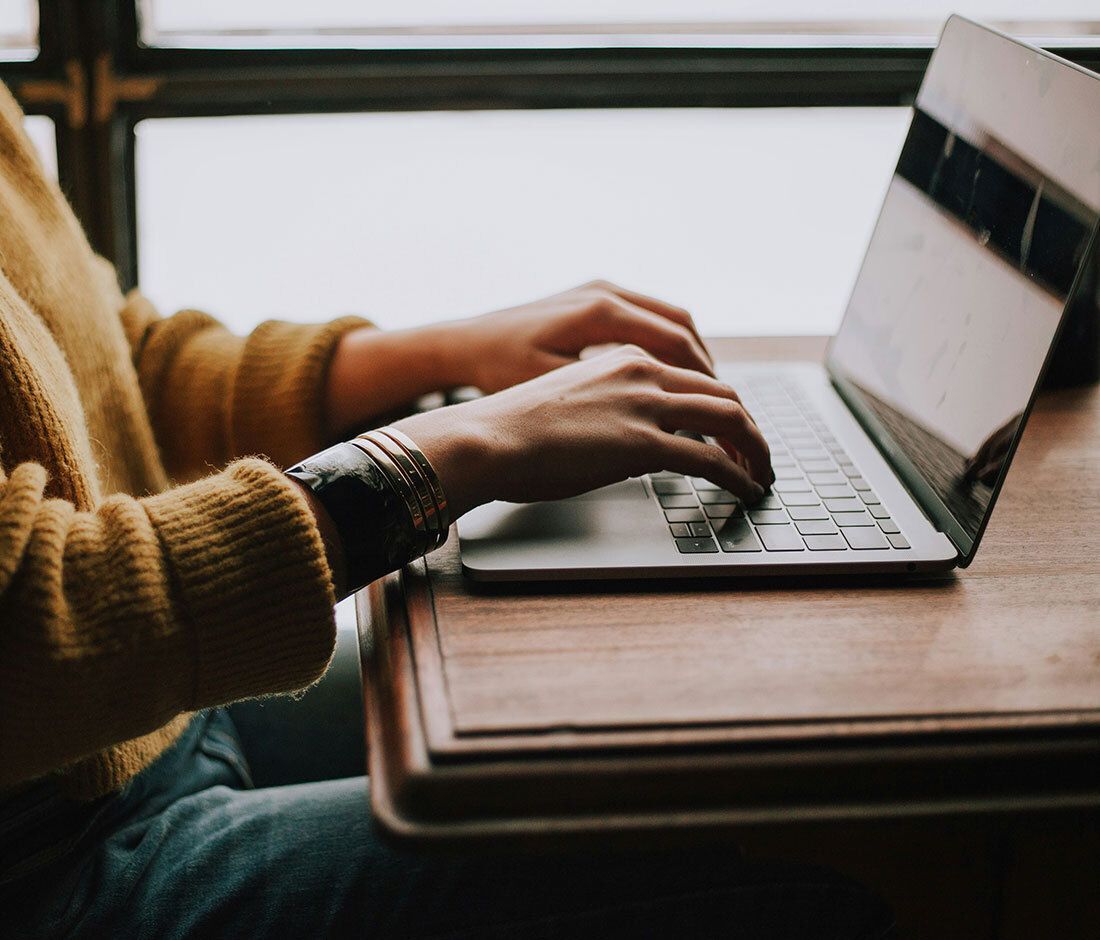 Person typing on a laptop at a wooden table, wearing a mustard sweater.