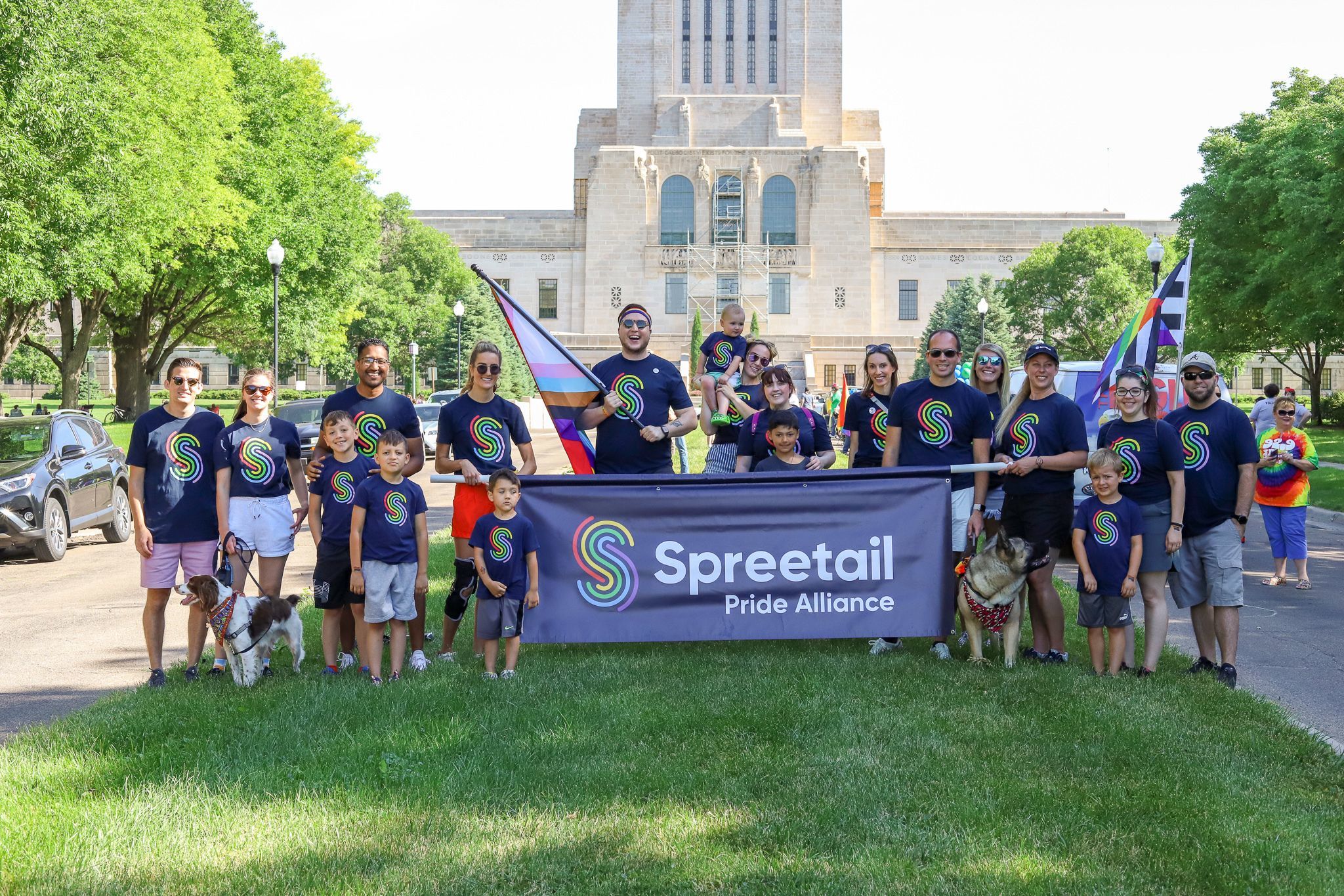 A group of Spreetail employees and their families pose in front of the Nebraska capitol with rainbow flags, rainbow t-shirts, and a banner that reads "Spreetail Pride Alliance."